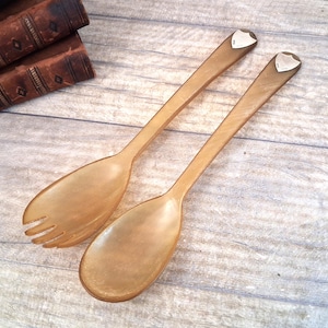 May include: A pair of salad servers made from translucent light brown horn material. Each handle features a small, triangular silver-colored metal shield inlay at the top. One server is a solid spoon, while the other is a three-pronged fork.