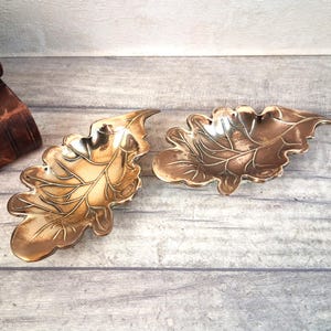 May include: Two metallic leaf-shaped dishes with a polished gold-tone finish. Each dish features embossed vein details and a curved, organic silhouette. The items are displayed side-by-side, showcasing a reflective surface and intricate edges.