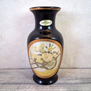 Könnte beinhalten: Schwarze Vase mit goldenem Rand und dekorativem Blumendesign. Die Vase hat ein ovales Gold-Weiß-Design mit Blumenarrangement und einem kleinen Aufkleber mit der Aufschrift "24KT GOLD".