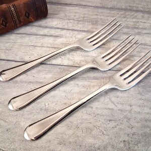May include: A set of three polished stainless steel dinner forks arranged diagonally. Each fork features a smooth, rounded handle and four tines. The metal surface reflects light, showing a bright, clean finish on the tines and the curved handles.