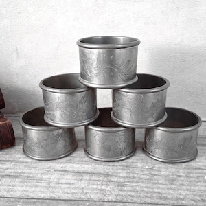 May include: A set of six cylindrical silver-toned metal napkin rings stacked in a pyramid formation. Each ring features a detailed engraved pattern of a dragon with visible scales and flowing tail elements wrapping around the exterior surface.