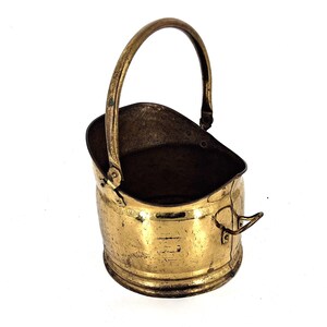 May include: A vintage brass bucket with a curved handle and a small side handle. The bucket has a rounded base and a slightly flared top. The brass has a warm, golden hue with signs of age and use.
