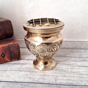 May include: A polished brass flower frog vase featuring a rounded body with etched leaf and floral patterns. The vessel includes a removable metal grid top designed for arranging stems, set on a sturdy pedestal base with a metallic gold finish.