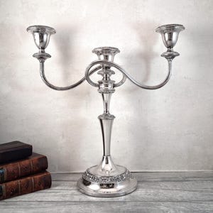 May include: A silver-toned metal candelabra featuring three candle holders with curved arms extending from a central stem. The base and the central column are decorated with embossed floral patterns, and the surface has a polished finish.