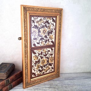 May include: An antique gold-framed artwork with a floral design. The artwork is divided into two panels, featuring a pattern in brown, tan, and cream. The frame has detailed carvings. The piece is leaning against a wall.