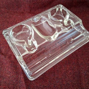 Op de afbeelding: Een bureau-organizer van helder glas met twee ronde pennenbakjes en een centraal rechthoekig vak. De basis is rechthoekig met een getrapt ontwerp, wat zorgt voor een stabiele ondergrond voor de geïntegreerde opbergvakken.