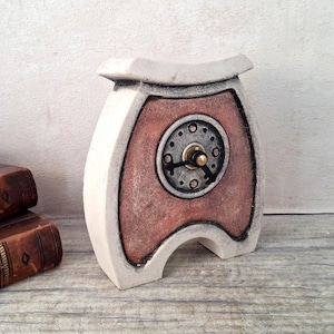 May include: A tabletop clock with a curved, off-white frame and a textured, terracotta-coloured centre panel. The clock face is circular, featuring a metallic grey finish with small circular indentations and black hands with a central gold-toned pin.