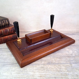 May include: A vintage wooden desk tidy with a dark brown leather-effect surface. It features a small wooden tray, two pen holders with black tops and gold bases, and a rectangular design. The organizer is surrounded by a stack of old books.