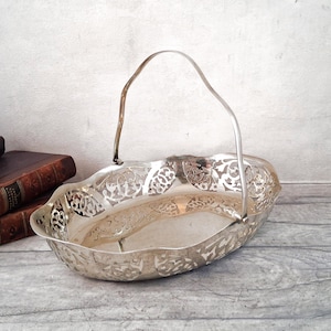 May include: An oval silver-toned metal basket featuring an intricate openwork filigree pattern along the sides, a scalloped rim, and a single arched handle attached to the sides. The basket has a polished, reflective surface finish.