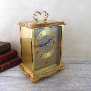 Könnte beinhalten: Eine goldfarbene Kaminuhr mit einem Glaszifferblatt und einem dekorativen Griff oben. Das Zifferblatt der Uhr hat römische Ziffern und die Worte "Stueben Quartz" sind darauf gedruckt. Die Uhr steht auf einer Holzoberfläche.