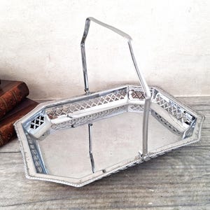 May include: A silver-toned metal serving tray with a rectangular shape and beveled corners. The tray features an intricate openwork lattice pattern along the raised sides and a single tall, arched handle attached to the centre of the long edges.