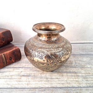 May include: A round brass pot featuring an intricate engraved pattern of animals and foliage. The vessel has a wide, flared rim and a polished metallic finish with darkened recessed areas that highlight the detailed etched designs on its surface.
