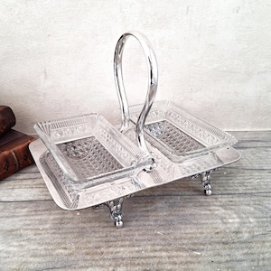 May include: A silver-plated serving stand featuring two rectangular clear glass dishes with a diamond-cut pattern. The metal base has four ornate feet and a central arched handle, providing a structured frame for the two removable glass containers.