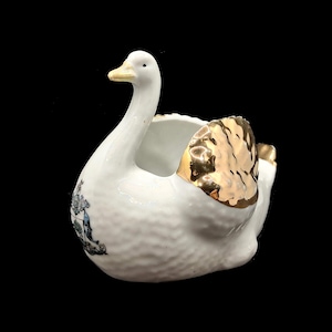 Pode incluir: Um recipiente em forma de cisne de cerâmica branca com uma asa e um bico dourados. O recipiente tem uma superfície texturizada e um pequeno desenho floral azul e verde na lateral. O cisne tem um pescoço longo e curvo.