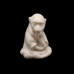 Peut inclure: Petite figurine de singe en céramique blanc cassé, assis, le menton posé sur la main. Le singe affiche une expression contemplative sur fond noir. La figurine est lisse et brillante.