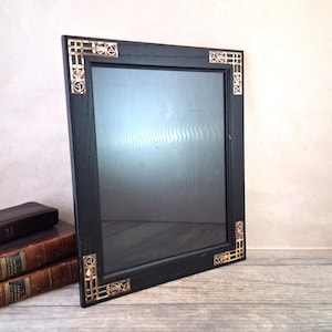 May include: A square black framed mirror with decorative gold corner accents. The mirror is leaning against a wall, with books in the background. The frame has a dark, matte finish. The mirror's dimensions are approximately 30 cm x 30 cm.