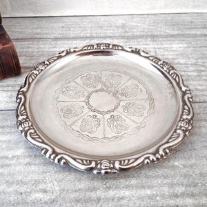 May include: A circular silver-coloured serving tray with a decorative, scalloped edge. The tray has an embossed floral pattern in the centre and around the rim. The tray is likely made of metal and is suitable for serving food or displaying decorative items.