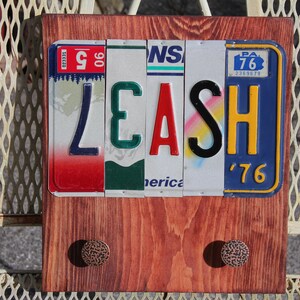 May include: Wooden leash holder with the word "LEASH" spelled out using repurposed license plates. The holder has two decorative knobs and is mounted on a wooden board. The license plates are in various colors, including red, green, and blue.