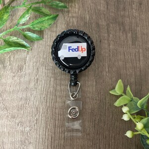 May include: A black badge reel with a bottle cap design. The badge reel features a white delivery truck graphic and the text "FedUp" in blue and red. The reel has a clear plastic strap and a metal clip.