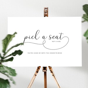 May include: A white sign on a wooden easel with the words "pick a seat" in elegant script. Below, it reads "NOT A SIDE" and "YOU'RE LOVED BY BOTH THE GROOM & BRIDE" in a simple font. Green foliage frames the sign.