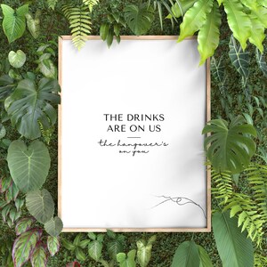 May include: A framed white print with the text "THE DRINKS ARE ON US" and "the hangover's on you" in black. The frame is surrounded by a variety of green plants and foliage, creating a natural, botanical theme.