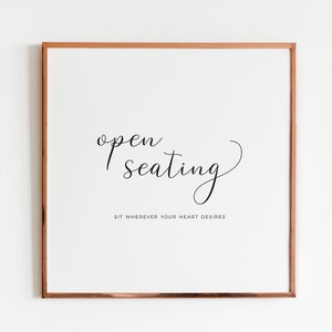 May include: Square framed print with the words "open seating" in elegant script. Below, the text reads "Sit wherever your heart desires." The frame is a warm copper color, and the background is white.