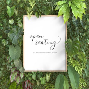 May include: A framed sign with the words "open seating" in elegant script, surrounded by lush green foliage. The sign also includes the text "Sit wherever your heart desires". The frame is a light wood color.