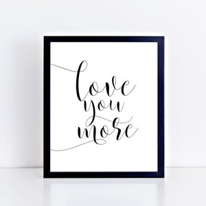 Puede incluir: Impresión enmarcada en blanco y negro con el texto "love you more" en escritura cursiva.