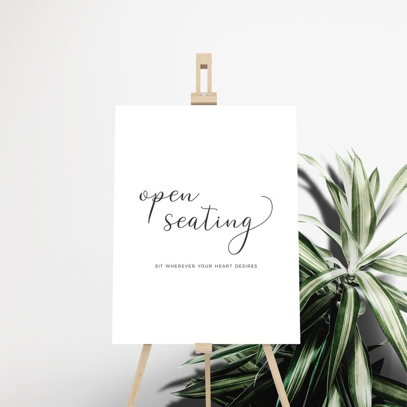 May include: A white sign on a wooden easel with the words "open seating" in elegant black script. Below, the text reads "SIT WHEREVER YOUR HEART DESIRES". A decorative plant is to the right.