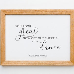 May include: A framed print with a light wood grain frame. The white background features elegant black script that reads "YOU LOOK great NOW GET OUT THERE & dance." Below, smaller text says "PLEASE HELP YOURSELF COMPLIMENTS OF THE NEWLYWEDS."