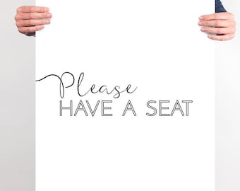 May include: A white poster with the text "Please HAVE A SEAT" in black. The word "Please" is written in a cursive font, while "HAVE A SEAT" is in a block font. The poster is held by a person.