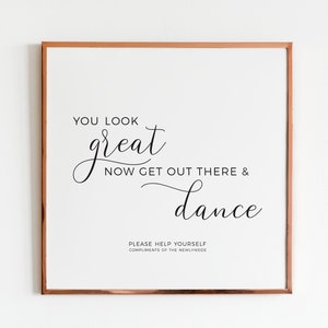 May include: A framed print with a copper-colored border. The white background features elegant black script that reads "YOU LOOK great NOW GET OUT THERE & dance." Below, it says "PLEASE HELP YOURSELF COMPLIMENTS OF THE NEWLYWEDS."