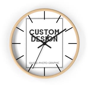May include: A white clock with black hands and a black square in the center. The square has the text "CUSTOM DESIGN" and "QUOTE-PHOTO-GRAPHIC" in black. The clock has a light wood frame.