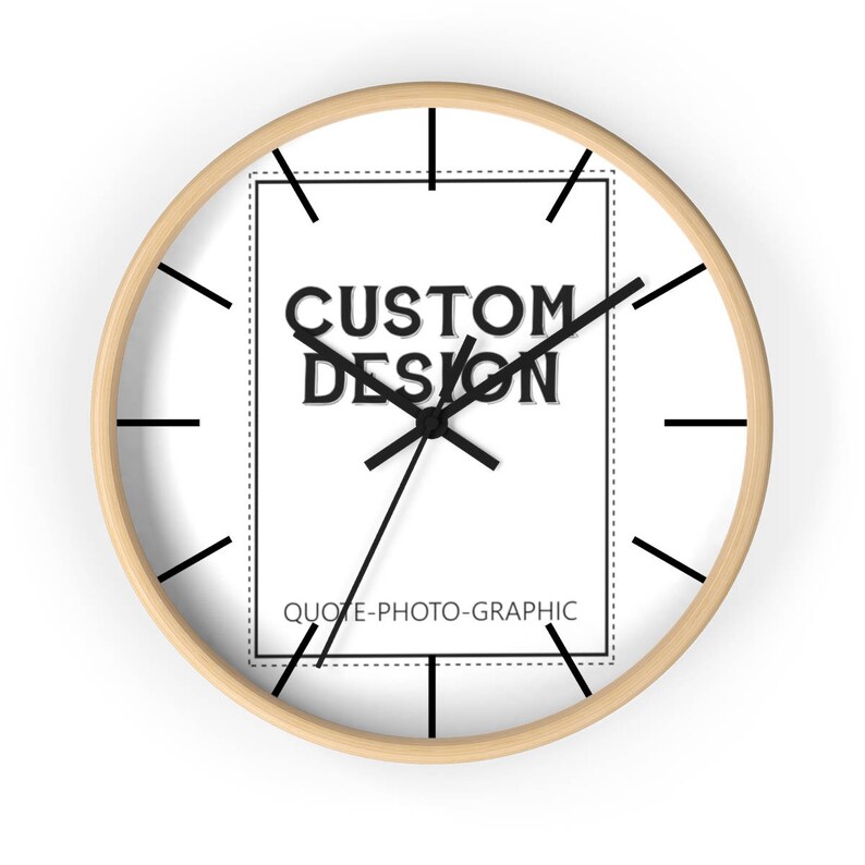 May include: A white clock with black hands and a black square in the center. The square has the text "CUSTOM DESIGN" and "QUOTE-PHOTO-GRAPHIC" in black. The clock has a light wood frame.