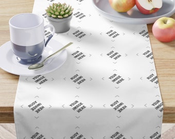 May include: White table runner with a repeating pattern of the text "YOUR IMAGE HERE" and "RUDY 30CM 383H". The runner is on a wooden table with a cup of coffee, a plate of apples, and a small potted succulent.