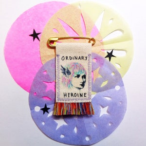 Peut inclure: Une épingle en tissu colorée avec un dessin d'une femme aux cheveux roses et les mots "ORDINARY HEROINE" dessus. L'épingle est fixée à une épingle de sûreté et est entourée de cercles de papier pastel avec des étoiles découpées.