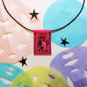 May include: A black cord necklace with a pink fabric pendant that says "CHANGE FOR NO ONE". The pendant has a black and white image of a person's face. The necklace is displayed on a white background with colorful paper cutouts.