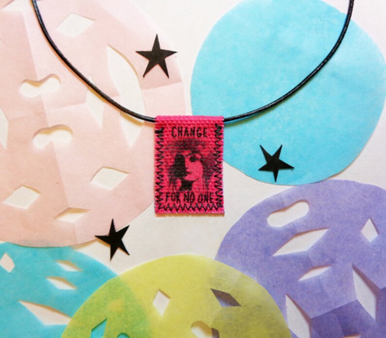 May include: A black cord necklace with a pink fabric pendant that says "CHANGE FOR NO ONE". The pendant has a black and white image of a person's face. The necklace is displayed on a white background with colorful paper cutouts.