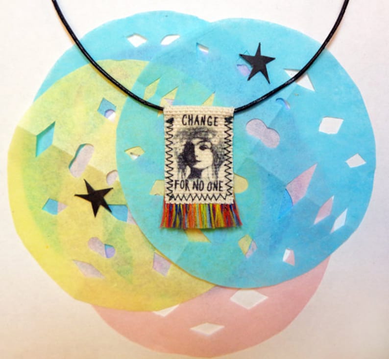 May include: A black leather cord necklace with a small fabric pendant. The pendant has a black and white image of a woman's face with the text "CHANGE FOR NO ONE" printed on it. The pendant has a fringe of colorful threads.