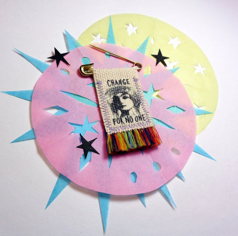 May include: A colorful fabric pin with a woman's face and the text "CHANGE FOR NO ONE". The pin is attached to a safety pin and is surrounded by pink and yellow paper with cut-out stars.