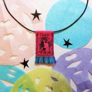 May include: A pink fabric pendant necklace with a black cord. The pendant has a fringe and says "CHANGE FOR NO ONE" with a black and white image of a woman's face.