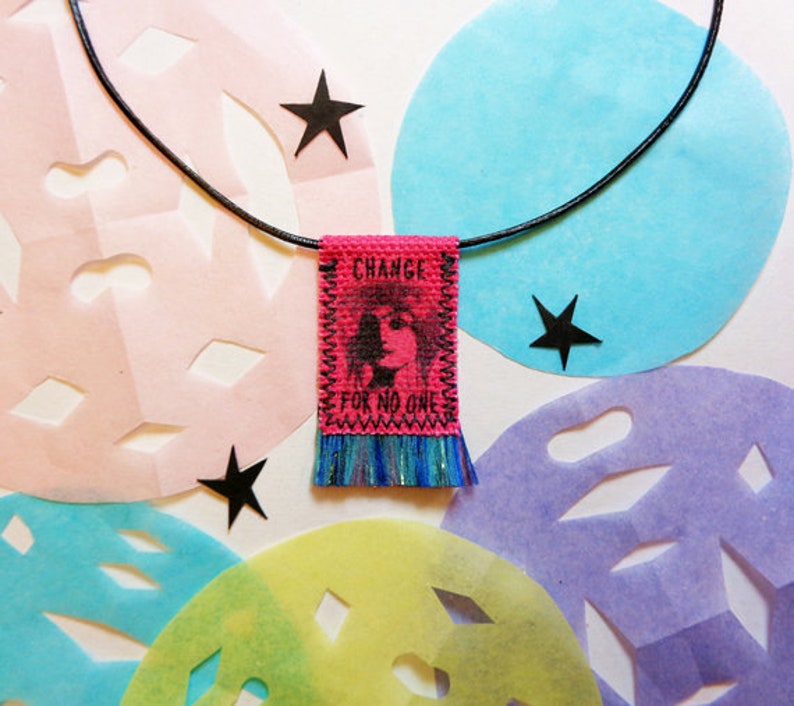 May include: A pink fabric pendant necklace with a black cord. The pendant has a fringe and says "CHANGE FOR NO ONE" with a black and white image of a woman's face.