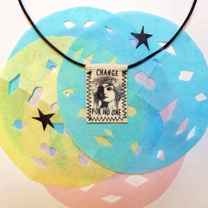 May include: A black cord necklace with a fabric tag that reads "CHANGE FOR NO ONE". The tag features a black and white illustration of a woman's face. The necklace is displayed on a background of blue, yellow, and pink paper with cut-out star shapes.