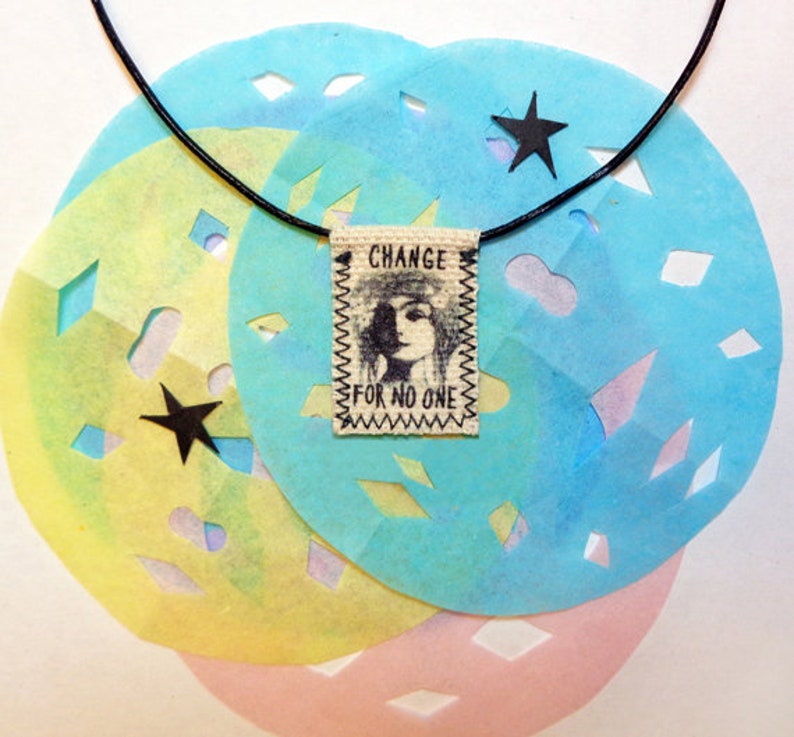 May include: A black cord necklace with a fabric tag that reads "CHANGE FOR NO ONE". The tag features a black and white illustration of a woman's face. The necklace is displayed on a background of blue, yellow, and pink paper with cut-out star shapes.