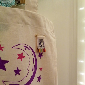 May include: A white canvas tote bag with a purple crescent moon and pink and purple stars printed on it. A small tag with the text "Change is for no one" is attached to the bag with a gold safety pin.