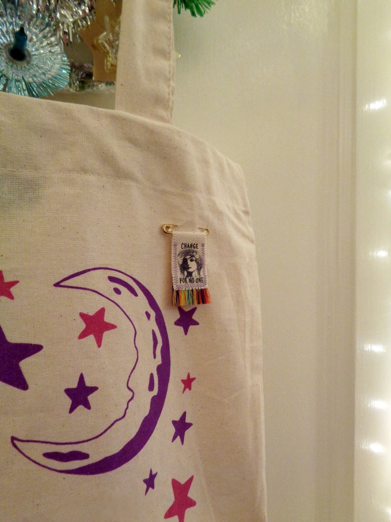 May include: A white canvas tote bag with a purple crescent moon and pink and purple stars printed on it. A small tag with the text "Change is for no one" is attached to the bag with a gold safety pin.