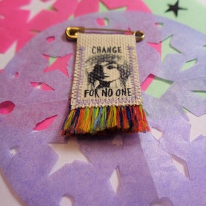 May include: A small fabric banner with a black and white illustration of a person's face and the text "CHANGE FOR NO ONE". The banner is attached to a gold safety pin and has a colorful fringe.