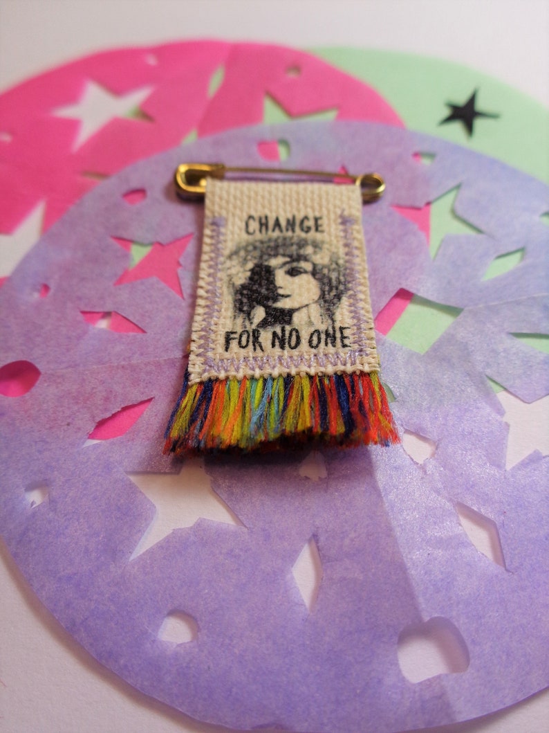 May include: A small fabric banner with a black and white illustration of a person's face and the text "CHANGE FOR NO ONE". The banner is attached to a gold safety pin and has a colorful fringe.