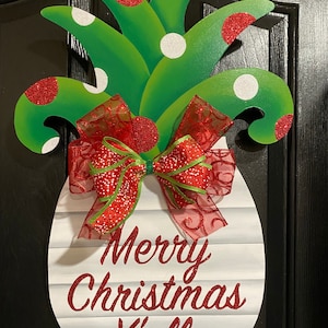 K&ouml;nnte beinhalten: Ein wei&szlig;er, ananasf&ouml;rmiger T&uuml;rkranz mit einer roten Schleife und dem Text "Merry Christmas Y'all". Die Ananas ist mit gr&uuml;nen Bl&auml;ttern, roten und wei&szlig;en Punkten verziert.