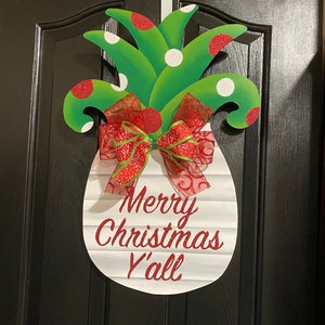 K&ouml;nnte beinhalten: Ein wei&szlig;er, ananasf&ouml;rmiger T&uuml;rkranz mit dem Text "Merry Christmas Y'all" in rotem Glitzer. Die Ananas hat gr&uuml;ne Bl&auml;tter mit roten und wei&szlig;en Punkten und eine rote Schleife mit gr&uuml;nem Besatz.